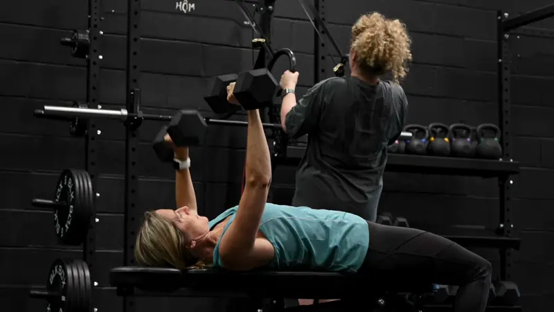 A woman dumbbell bench pressing while a Hōm coach spots her